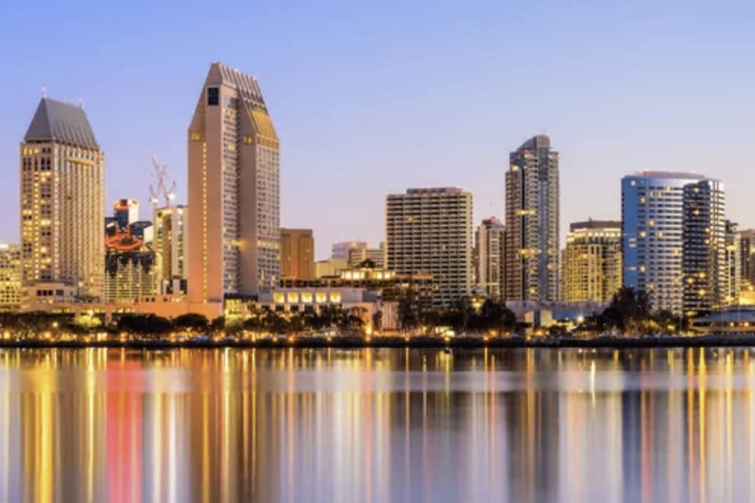 The skyline of a city with tall buildings at dusk, mirrored beautifully in a tranquil body of water, reminiscent of the elegant settings of previous conferences.