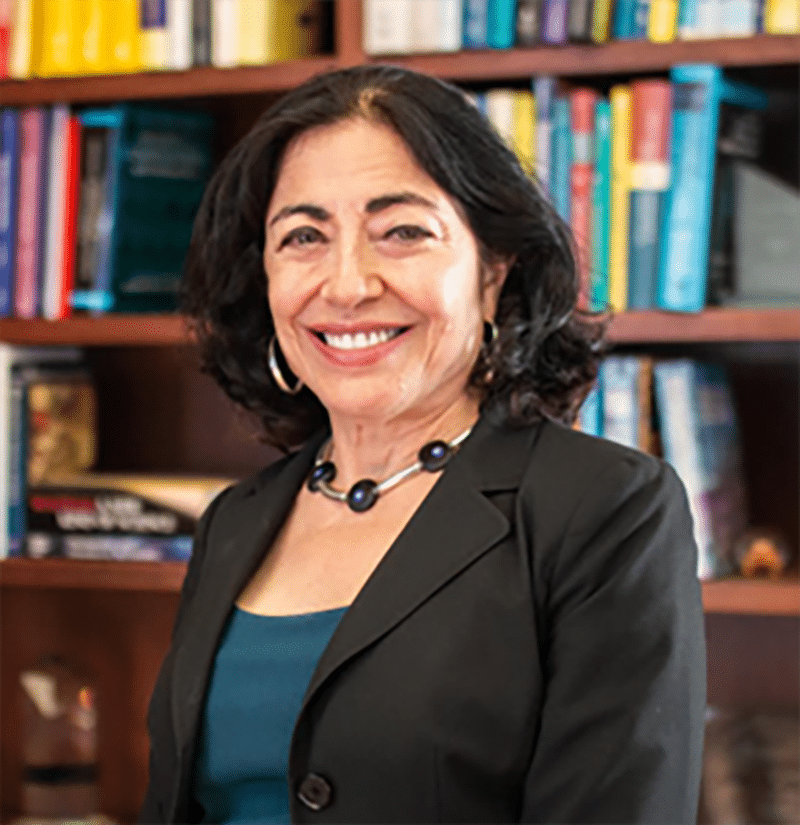 Jennifer Chayes, a woman with dark hair in a blazer and necklace, stands smiling in front of a bookshelf filled with books.