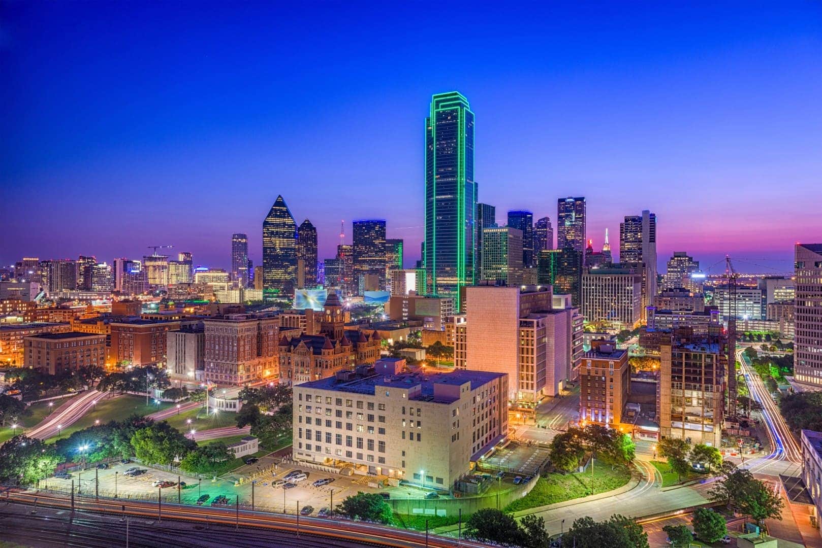 Downtown Dallas skyline at dusk with illuminated buildings, including a tall tower outlined in green lights, and city streets visible in the foreground.